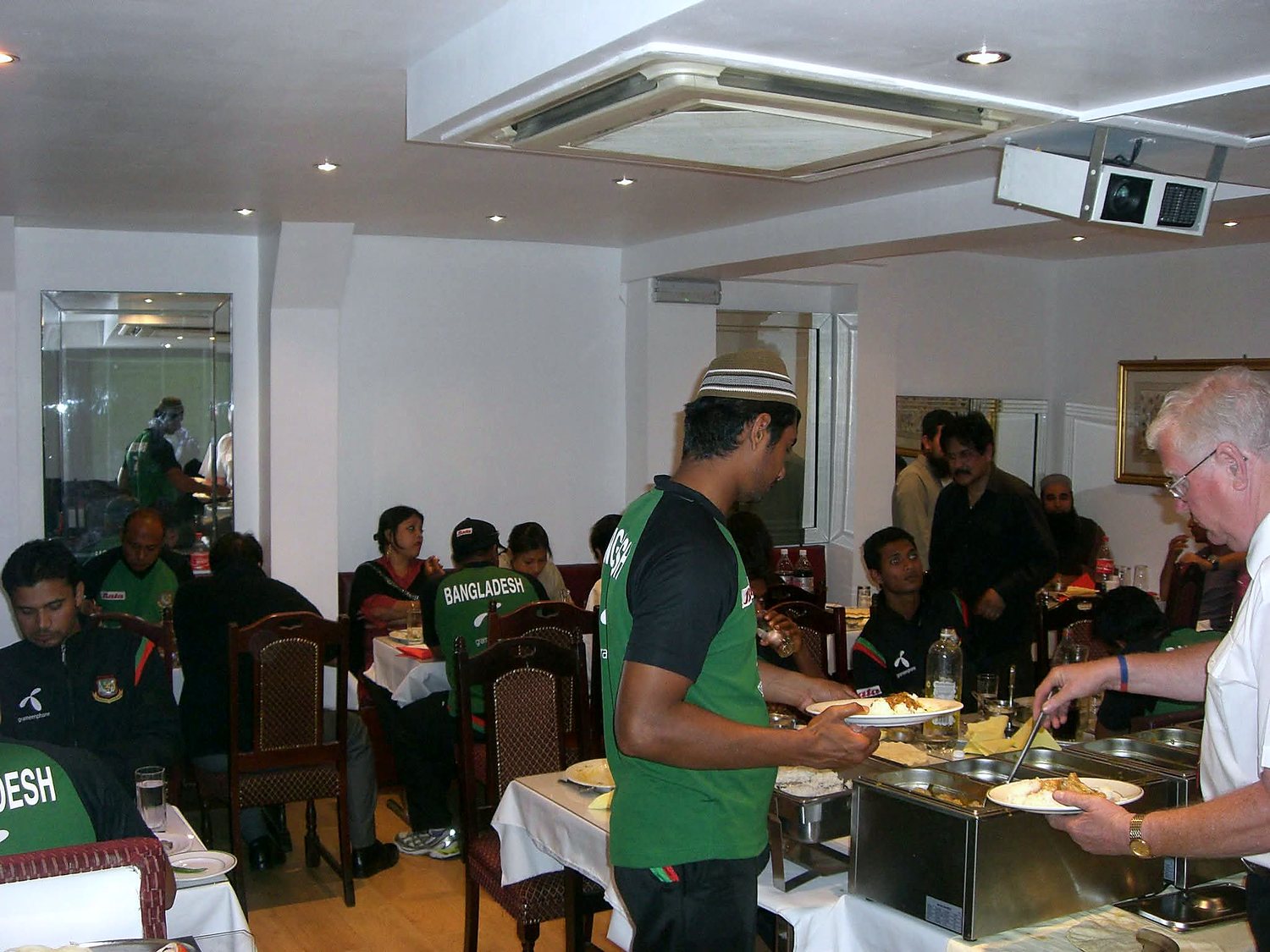 Bangladesh cricketers in green training kit filling plates from a buffet in the Balti House dining room, staff serving alongside them