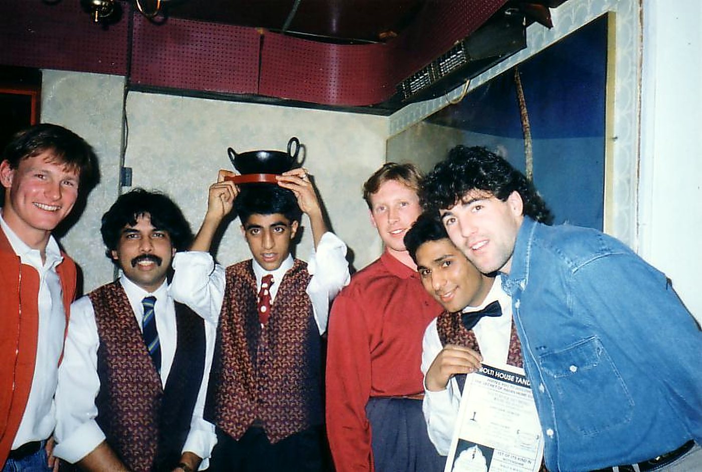 Six men in the Balti House in 1991 — three staff in patterned waistcoats, one balancing a black balti dish on a waiter’s head, and three guests