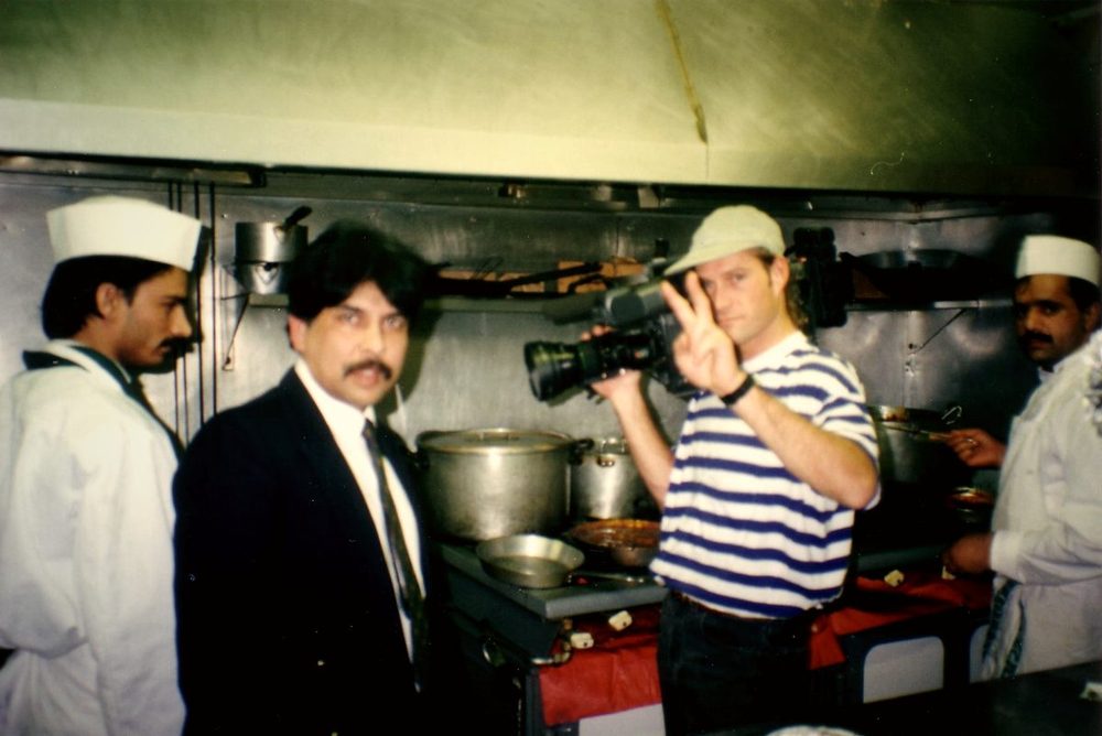 Sabbir Hossain in a suit beside a cameraman and chefs in the Balti House kitchen, 1995
