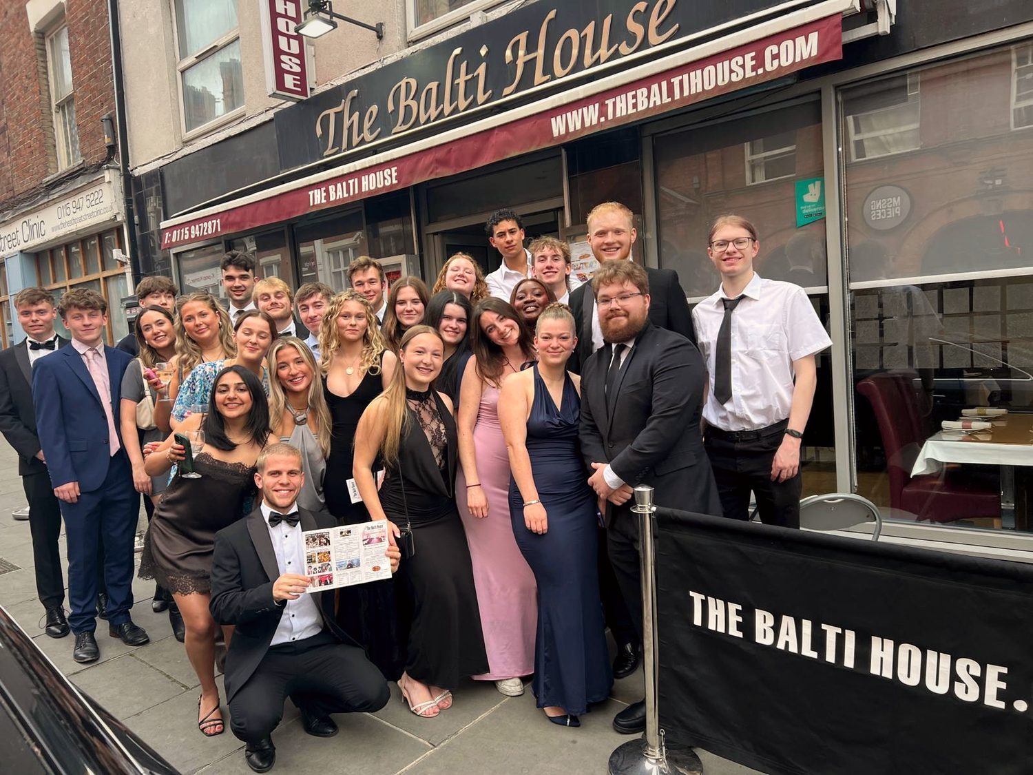 A large group in prom dresses and black tie posing outside The Balti House on Heathcoat Street, summer 2025